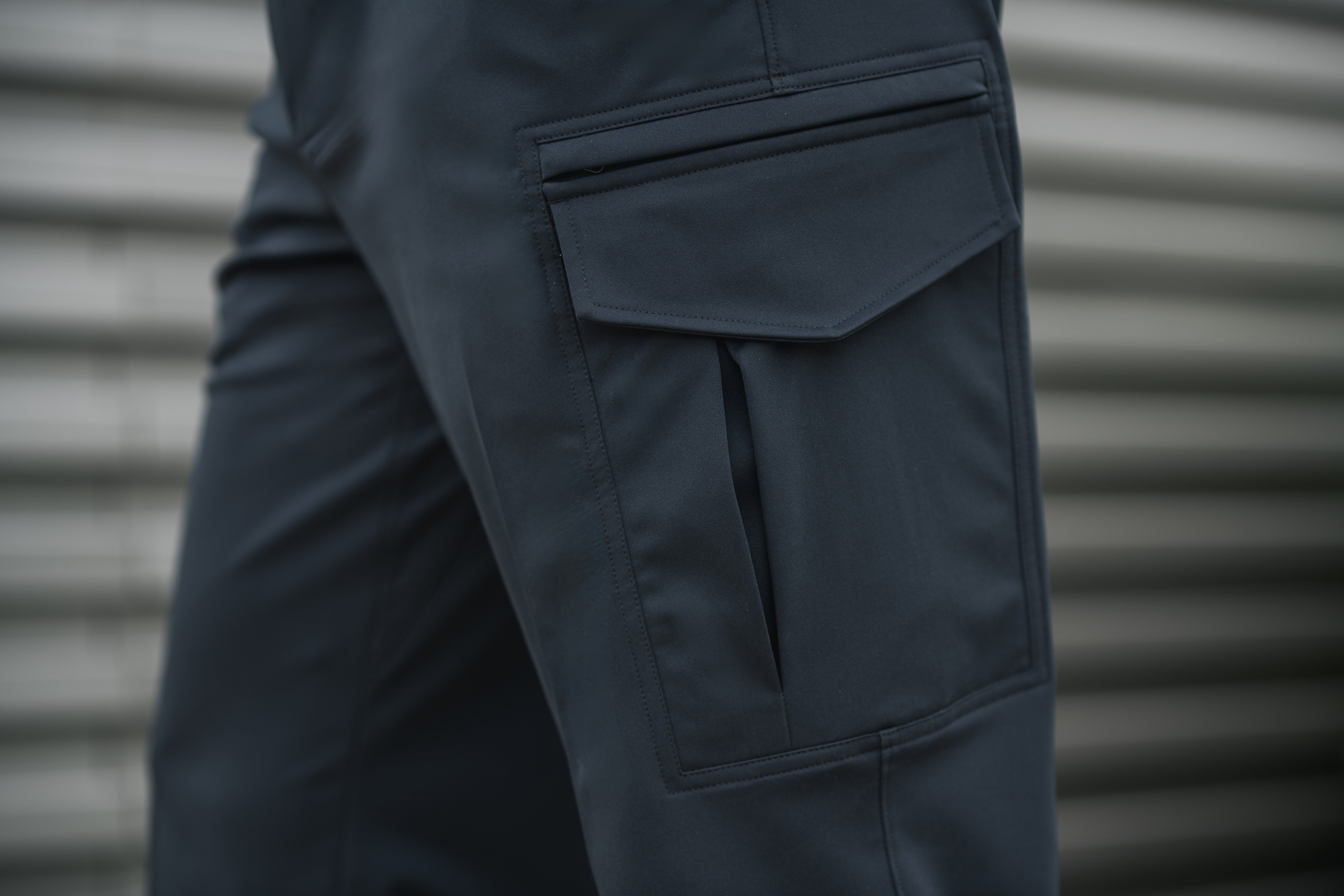 Photograph showing a close-up detail of dark charcoal gray cargo pants featuring a large, meticulously stitched pocket against a blurred background of corrugated metal siding showing subtle texture and color variations