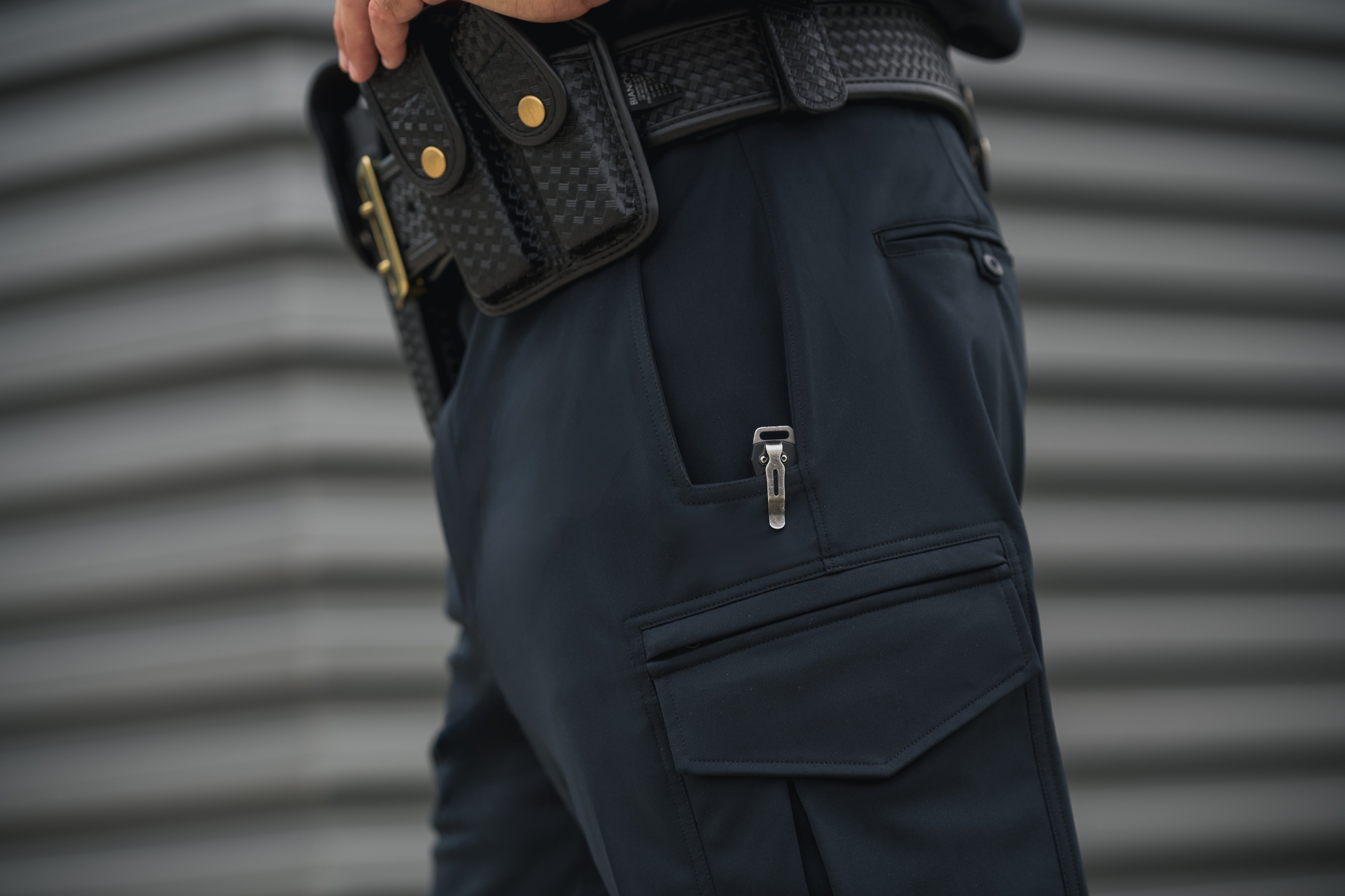 Photograph detail of a person wearing dark tactical pants with a utility pocket showing a silver folding knife against a gray corrugated metal background featuring a black woven belt with gold accents