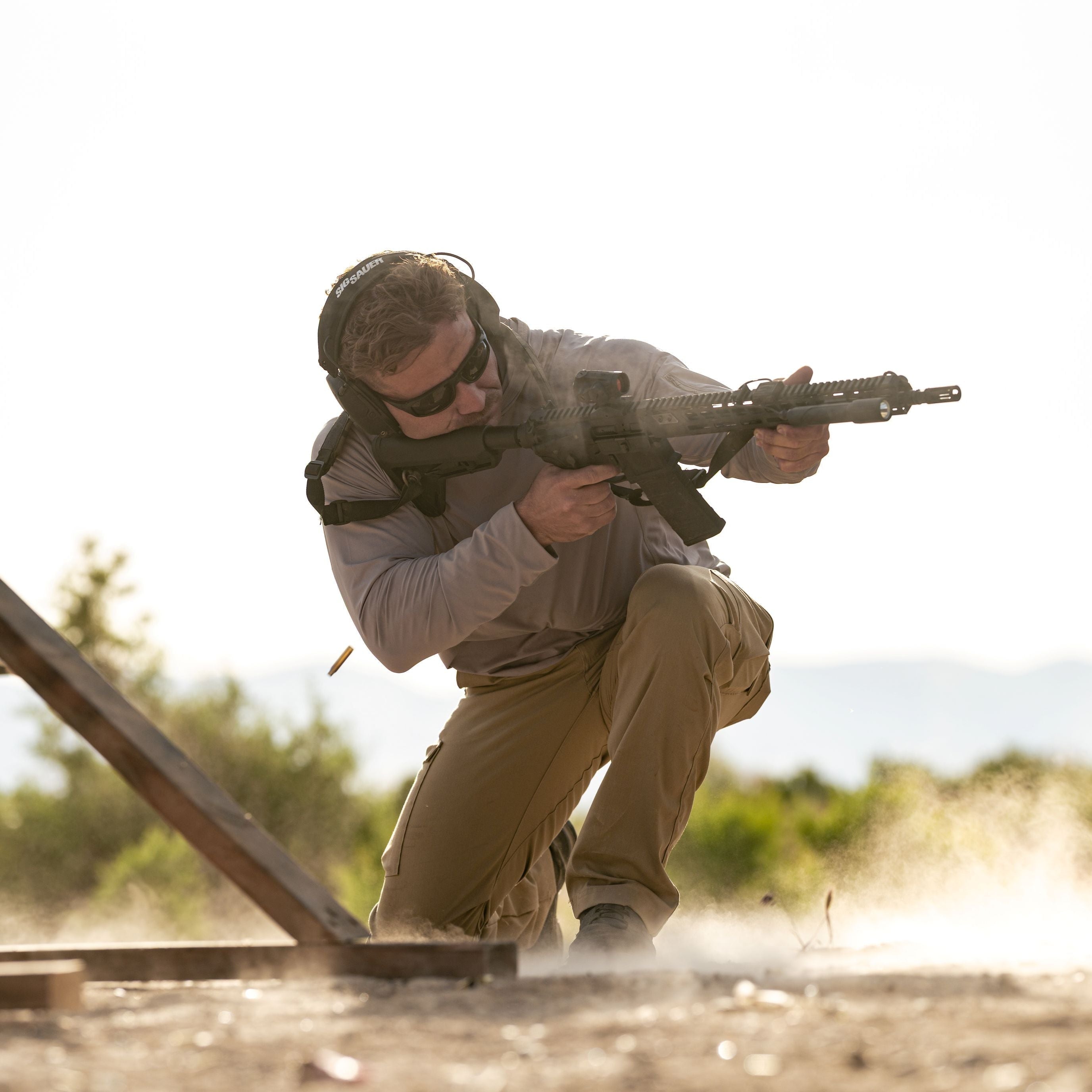 Photograph of a man in tan tactical gear firing a rifle at an outdoor range featuring a wooden structure and desert landscape with dust and smoke effects from the gunshot