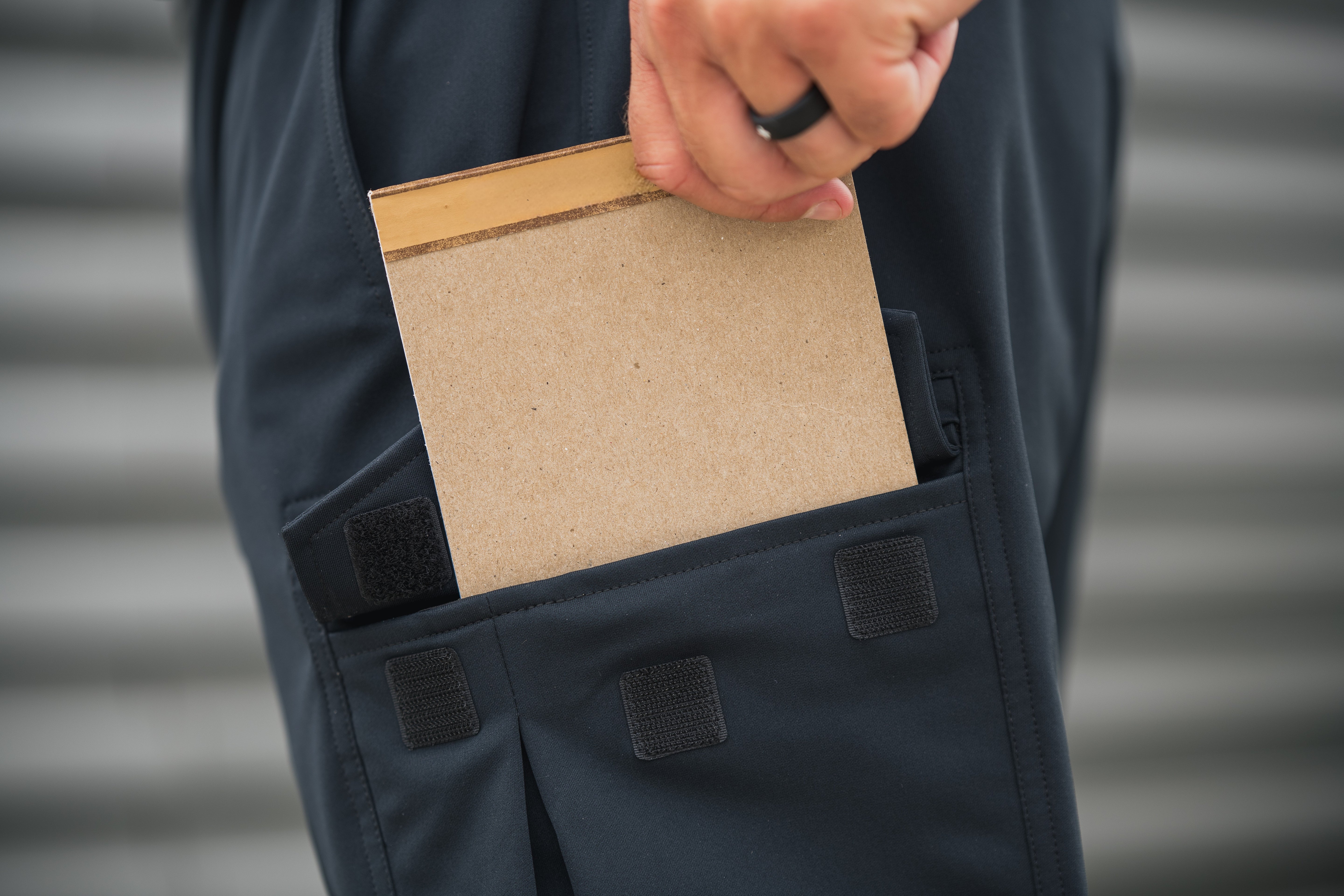 A photograph shows a person holding a blank, light brown cardboard notepad with a tan and brown trim in a dark-colored pocket against a blurred gray background
