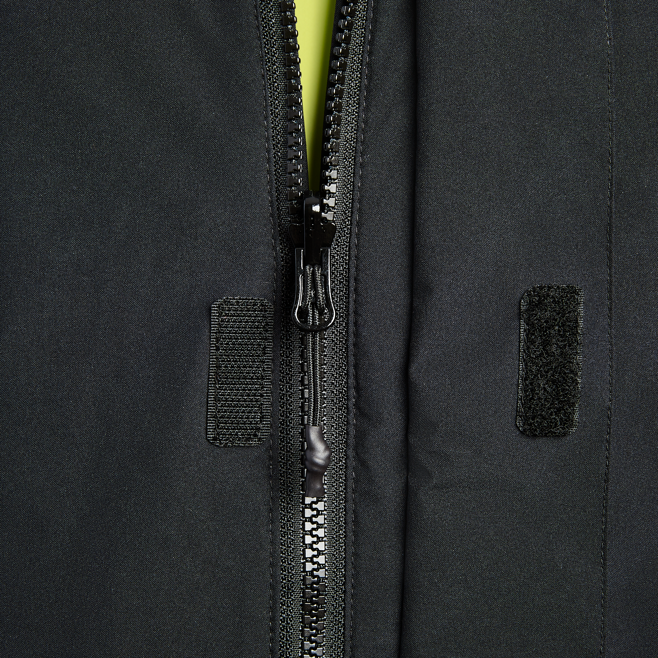 Close-up photograph of a black zipper and fabric texture featuring a shiny zipper pull, small black velcro patches, and subtle textural details.