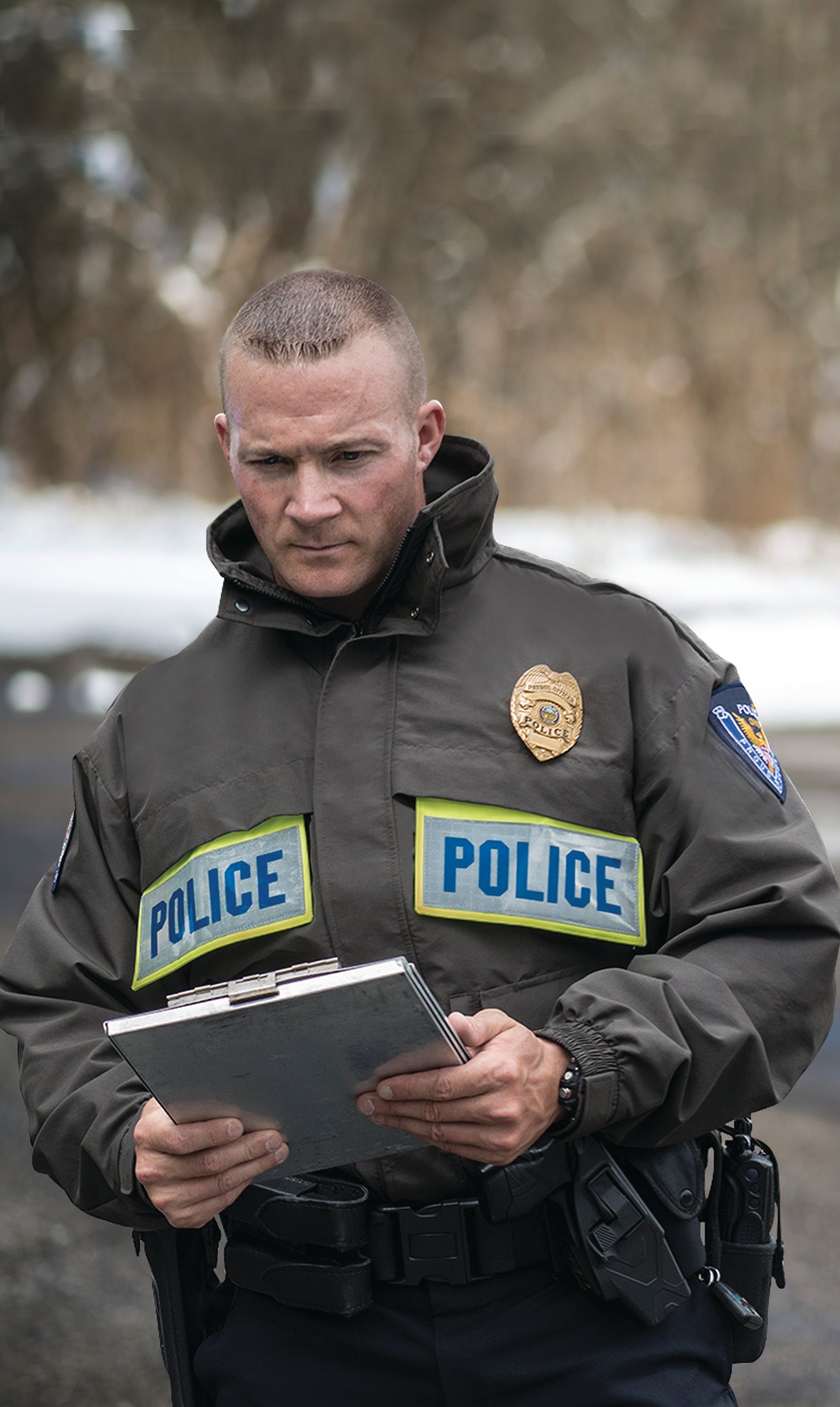 Photograph of a police officer outdoors reviewing notes on a clipboard in a snowy setting, wearing a dark grey jacket with POLICE patches, a gold badge, and a black utility belt with various tools.