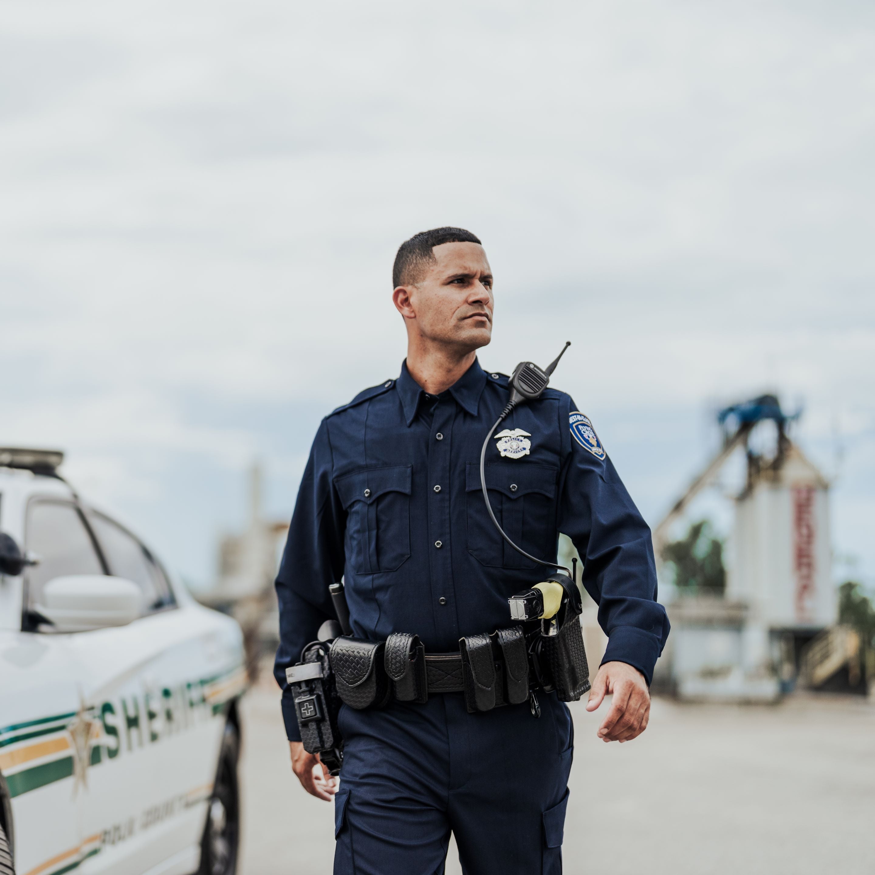 Photograph of a male sheriff in a dark blue uniform and duty belt near a sheriff's vehicle and industrial structures, featuring a radio, a badge, and gold-and-green vehicle stripes