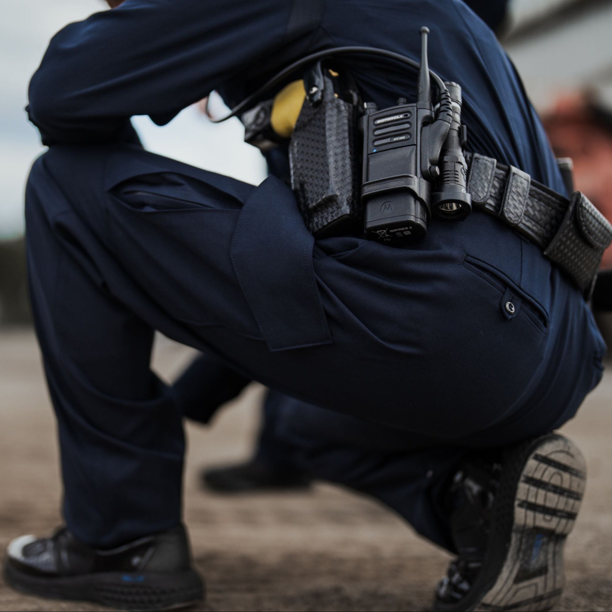 Photograph showing a police officer crouching outdoors with a Motorola APX radio, a flashlight, and a textured carbon fiber-like holster attached to their dark blue uniform and belt