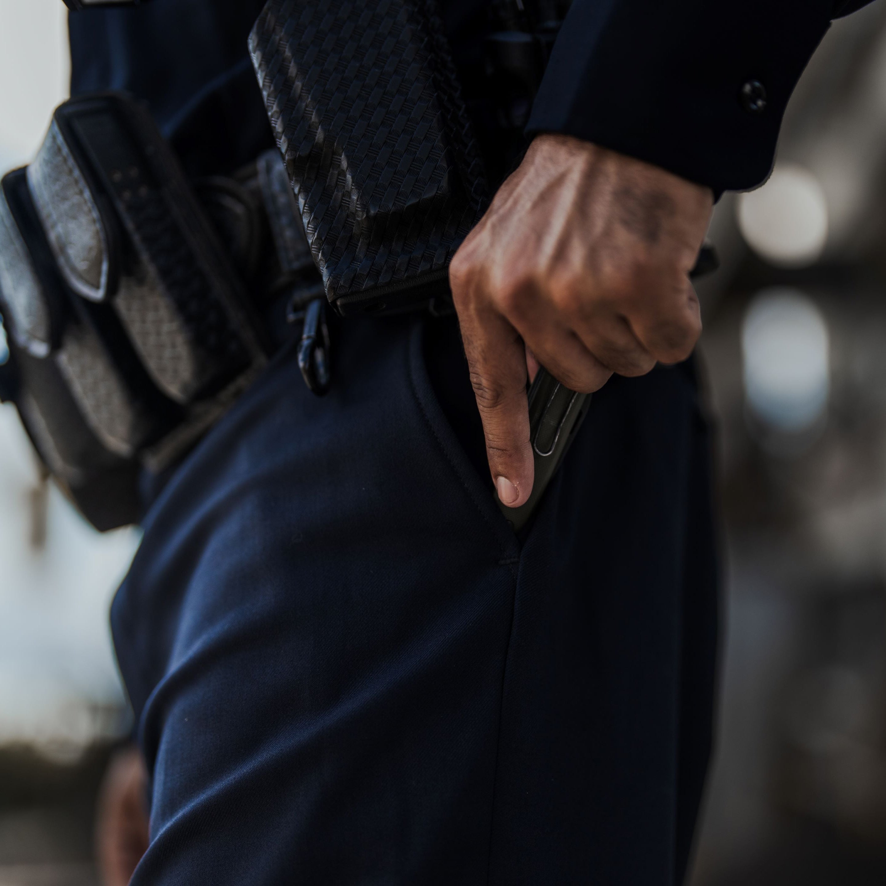 Photograph of a police officer's hand drawing a gun from his pocket outdoors with a dark blue uniform a textured black holster and a silver badge visible