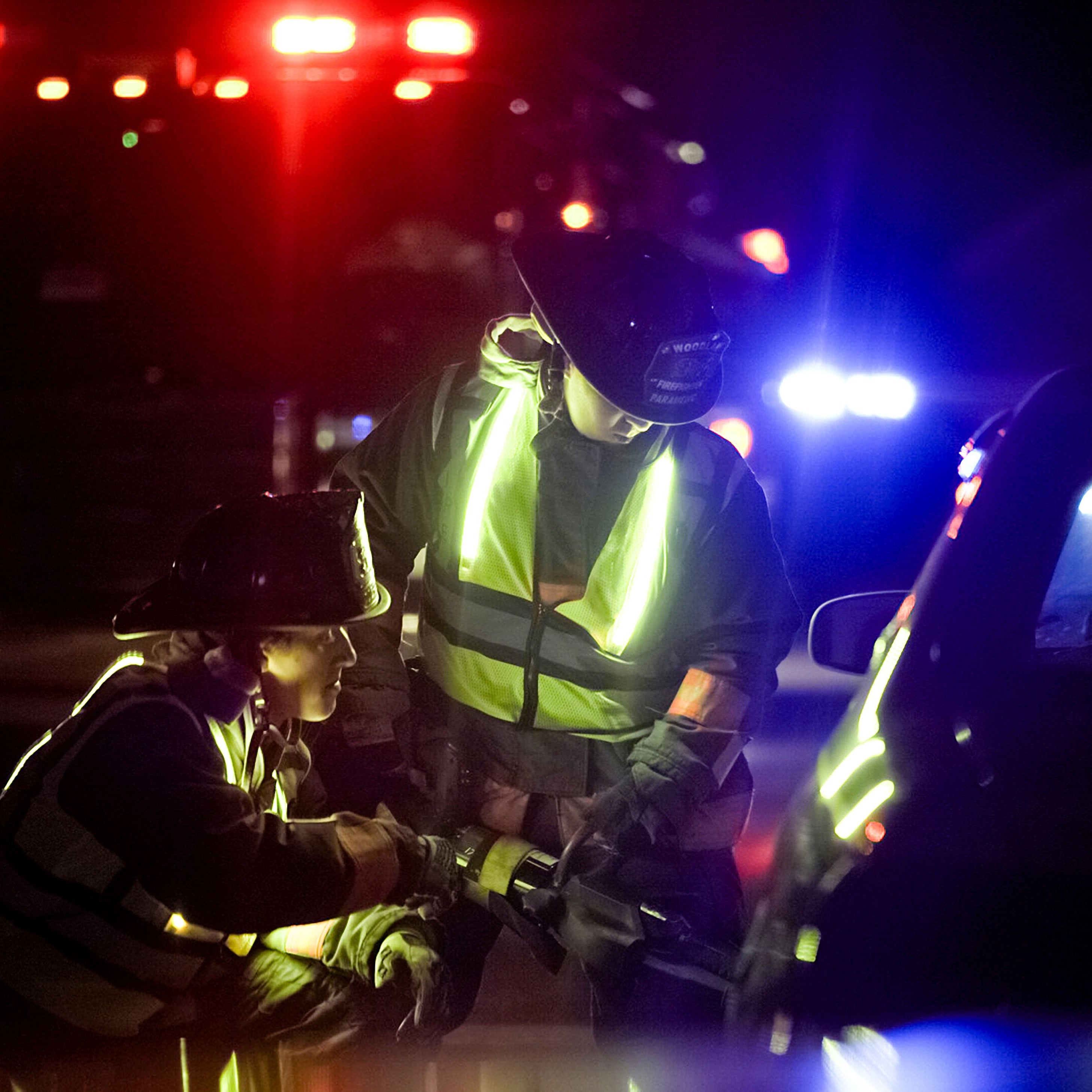 Nighttime photograph of firefighters in bright yellow vests using rescue tools at a car accident scene illuminated by red and blue emergency lights