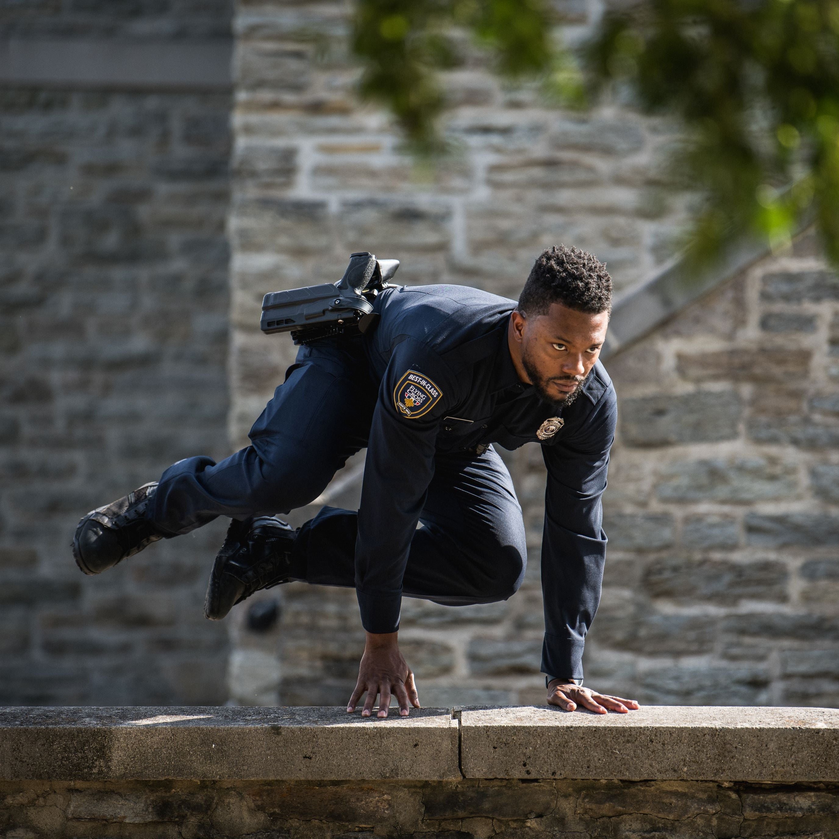 Photograph of a police officer in dynamic motion, leaping over a low stone wall near a historic stone building with a visible patch of green foliage and sunlit textures on the wall and officer's dark uniform.