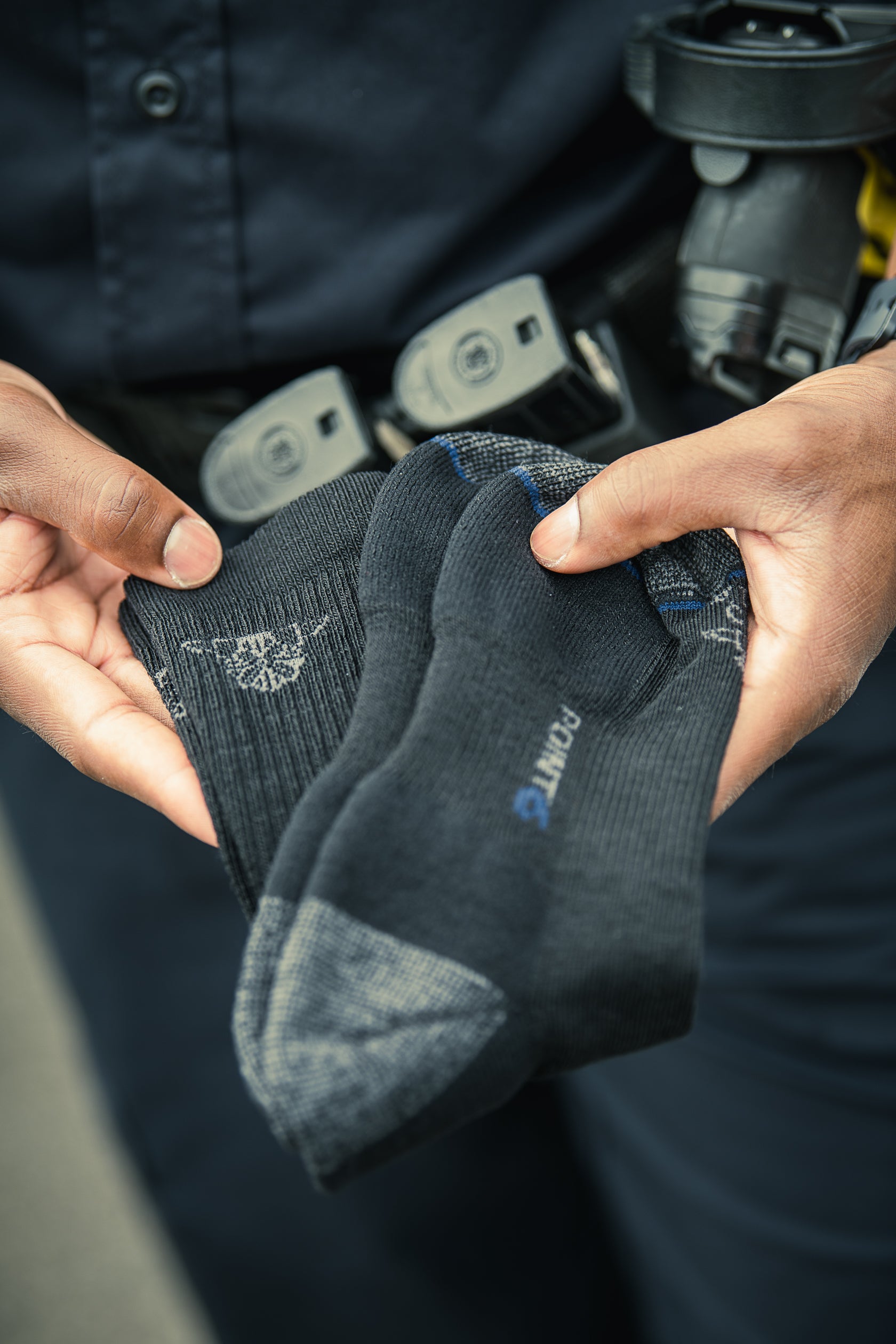 Photograph shows a person holding a pair of dark gray Point6 socks with a subtle pattern and blue detailing against a background of a dark uniform and holstered firearm