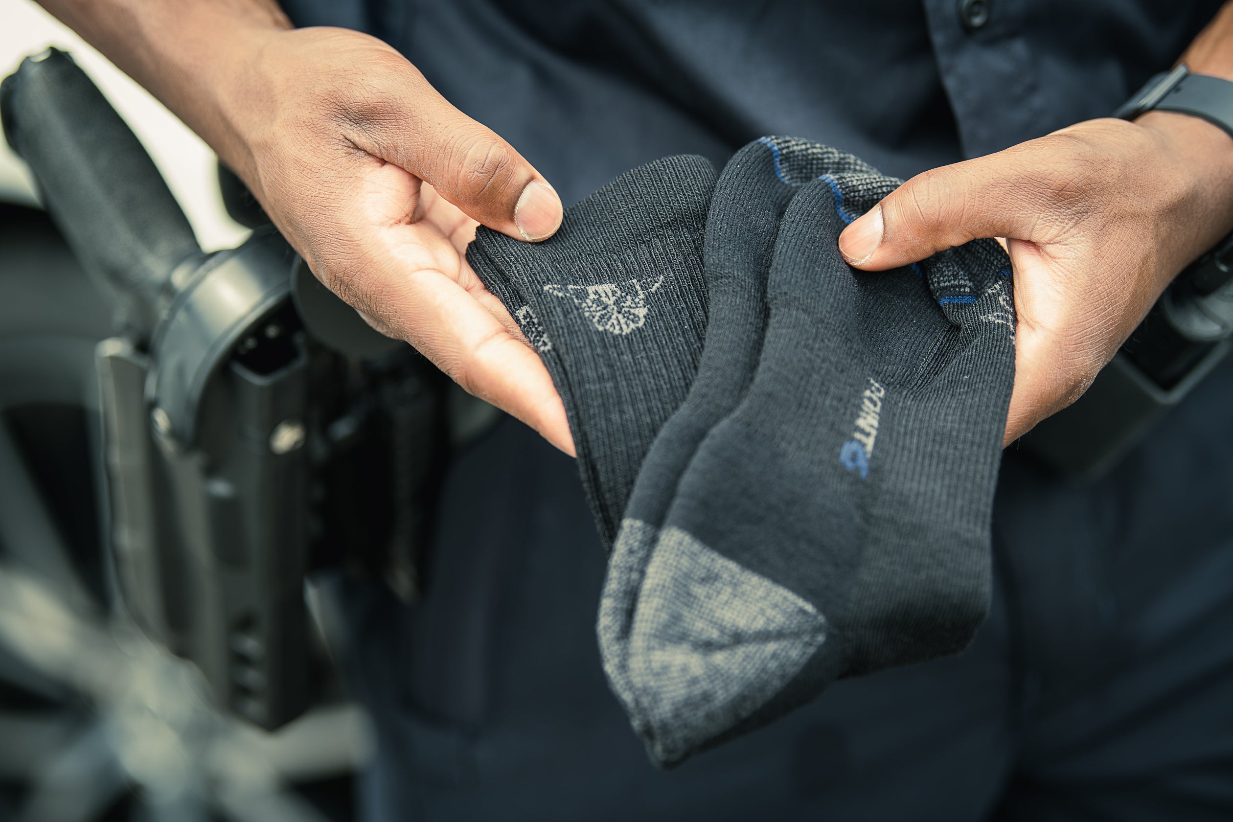 Photograph of a police officer's hands holding dark gray socks with a small logo and blue trim detail near a police car and holster.