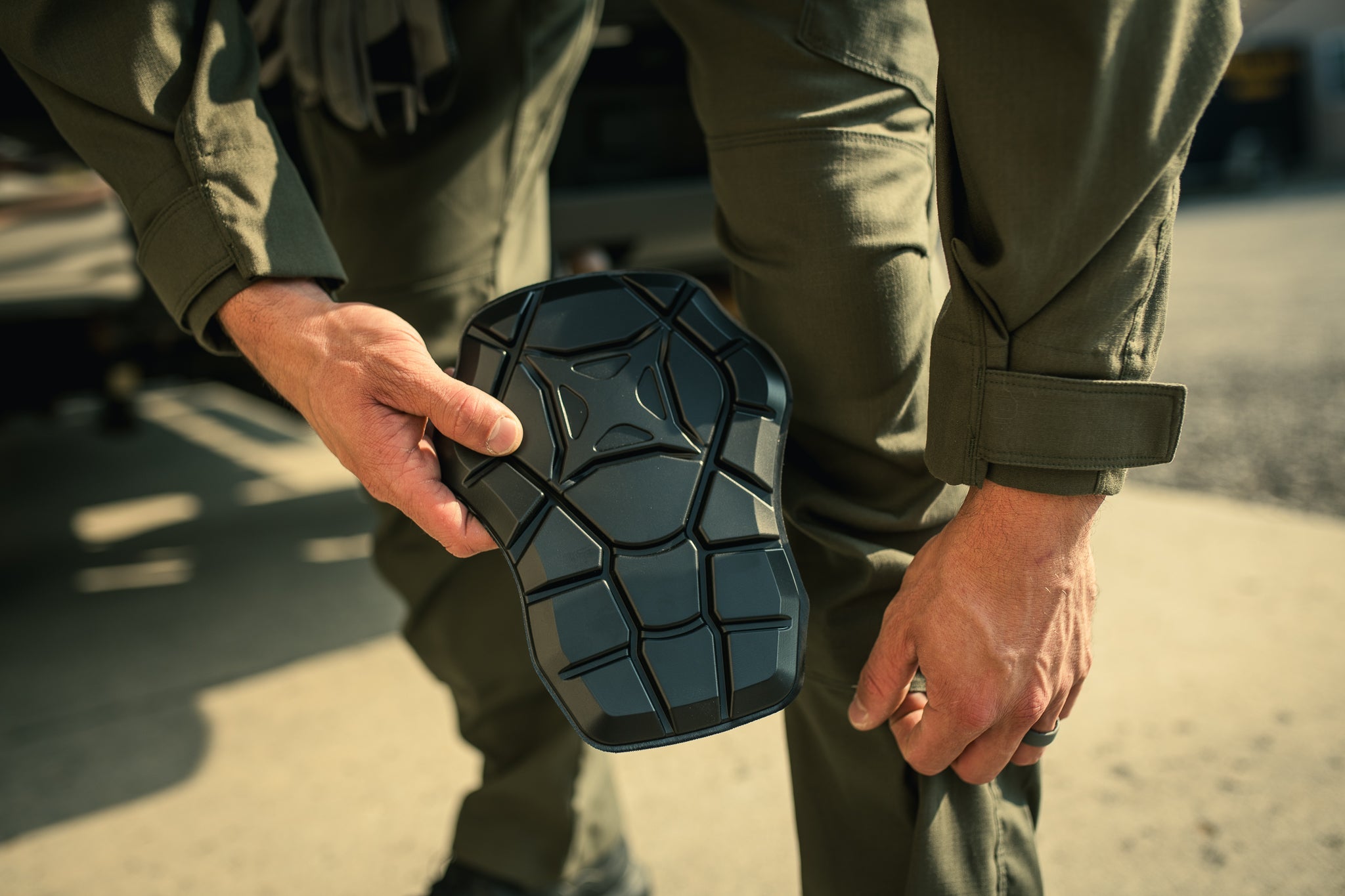 Photograph showing a person in olive green tactical pants holding a black, textured, knee pad with a geometric pattern outdoors on a sunlit asphalt surface