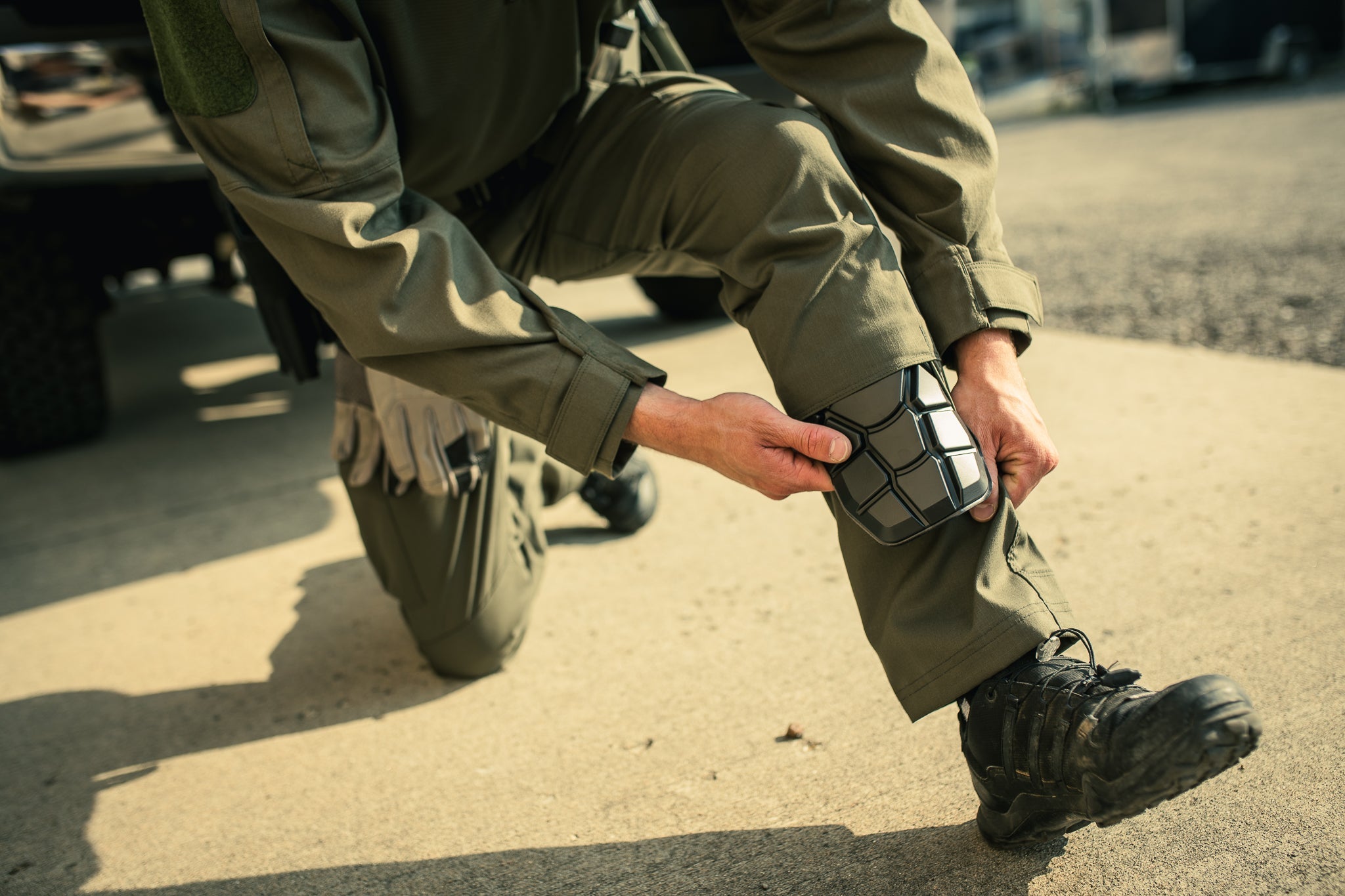 Photograph of a person in olive green tactical pants attaching black hexagonal knee pads outdoors near a vehicle showing textured fabric and sturdy footwear.