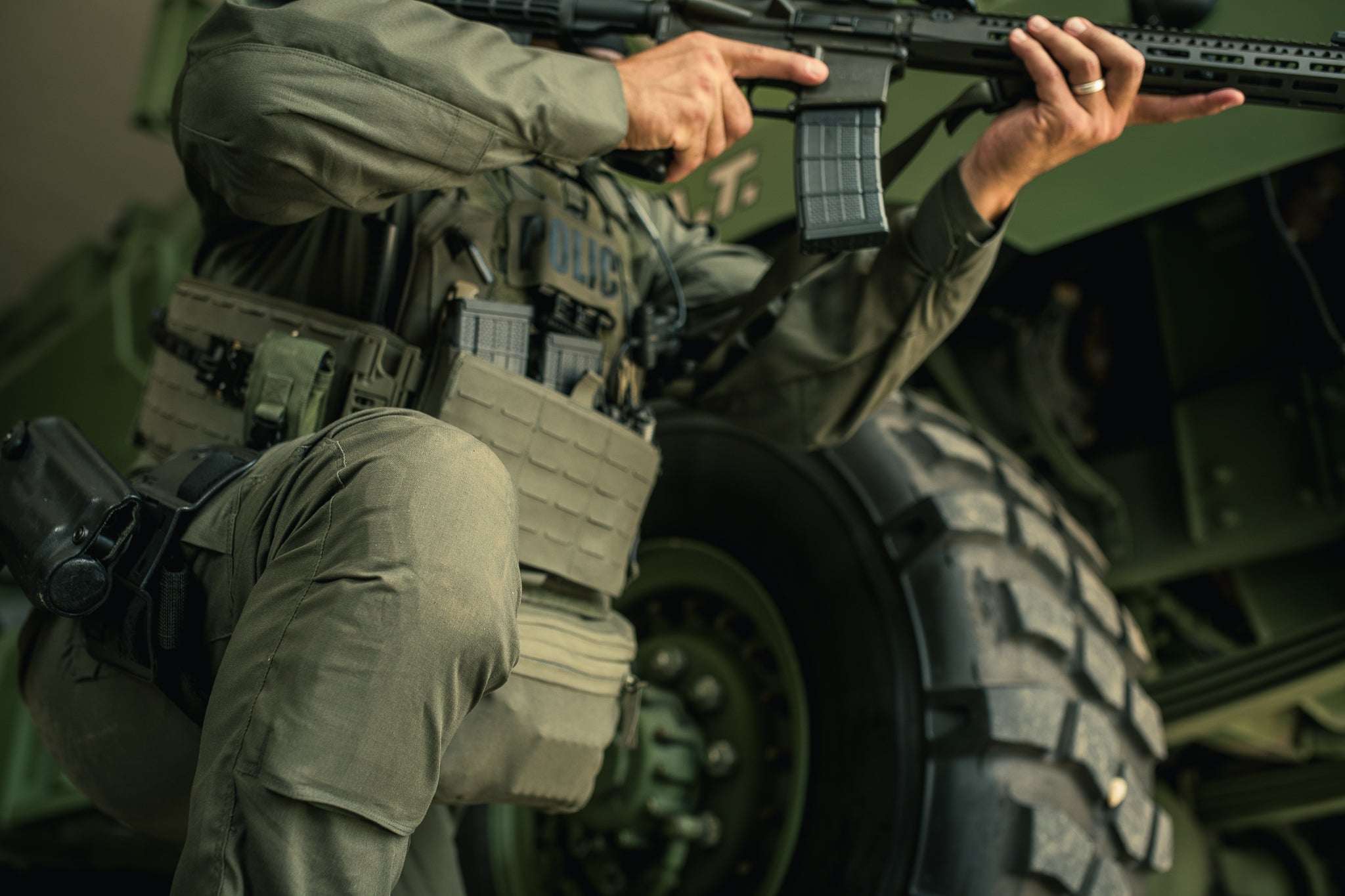 Photograph showing a police officer in tactical gear near a military vehicle with large tires and a visible POLICE patch on his vest his hands gripping a rifle showcasing a wedding band and olive drab uniform
