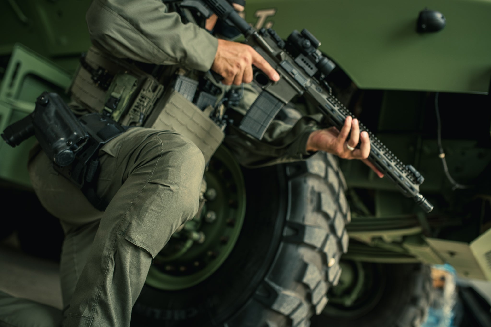 A photograph showing a soldier kneeling next to a military vehicle with a large tire and tactical gear including a rifle with scope and a sidearm in a holster, featuring olive green tones and textured fabric details