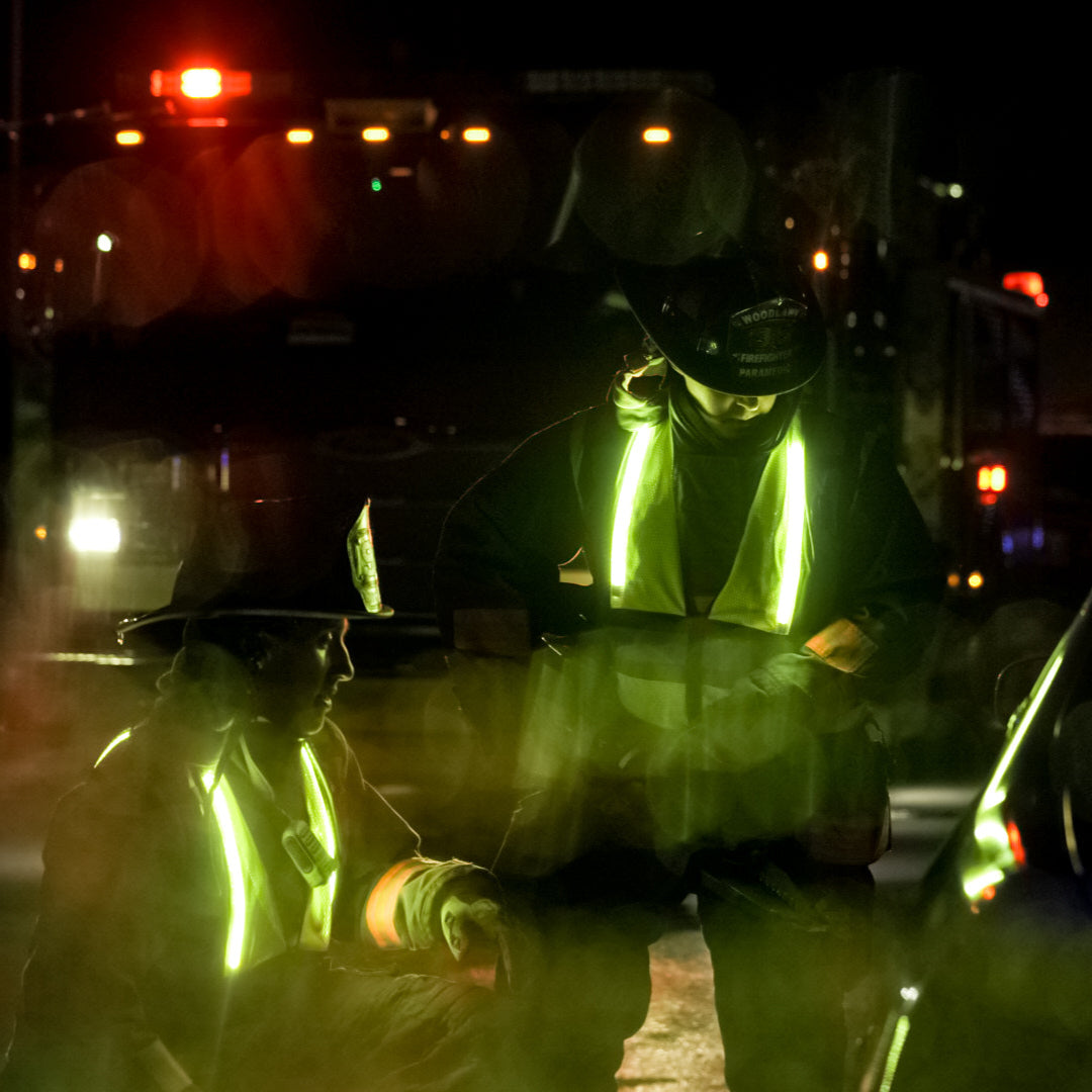 Night photography showing two firefighters in bright lime green safety vests at a nighttime emergency scene near a firetruck with flashing red and amber lights, one firefighter appears to be tending to an injury with a bandaged arm, the other is holding what looks like a medical bag