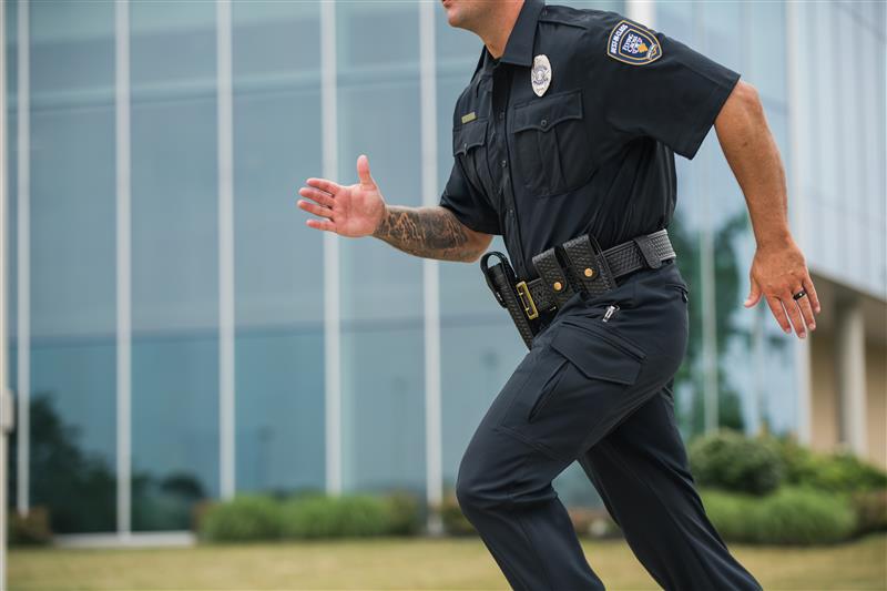 Photograph of a police officer running near a modern glass building showcasing a detailed view of his tactical gear including a black belt brass buckle and a visible tattoo a dark ring and cargo pants