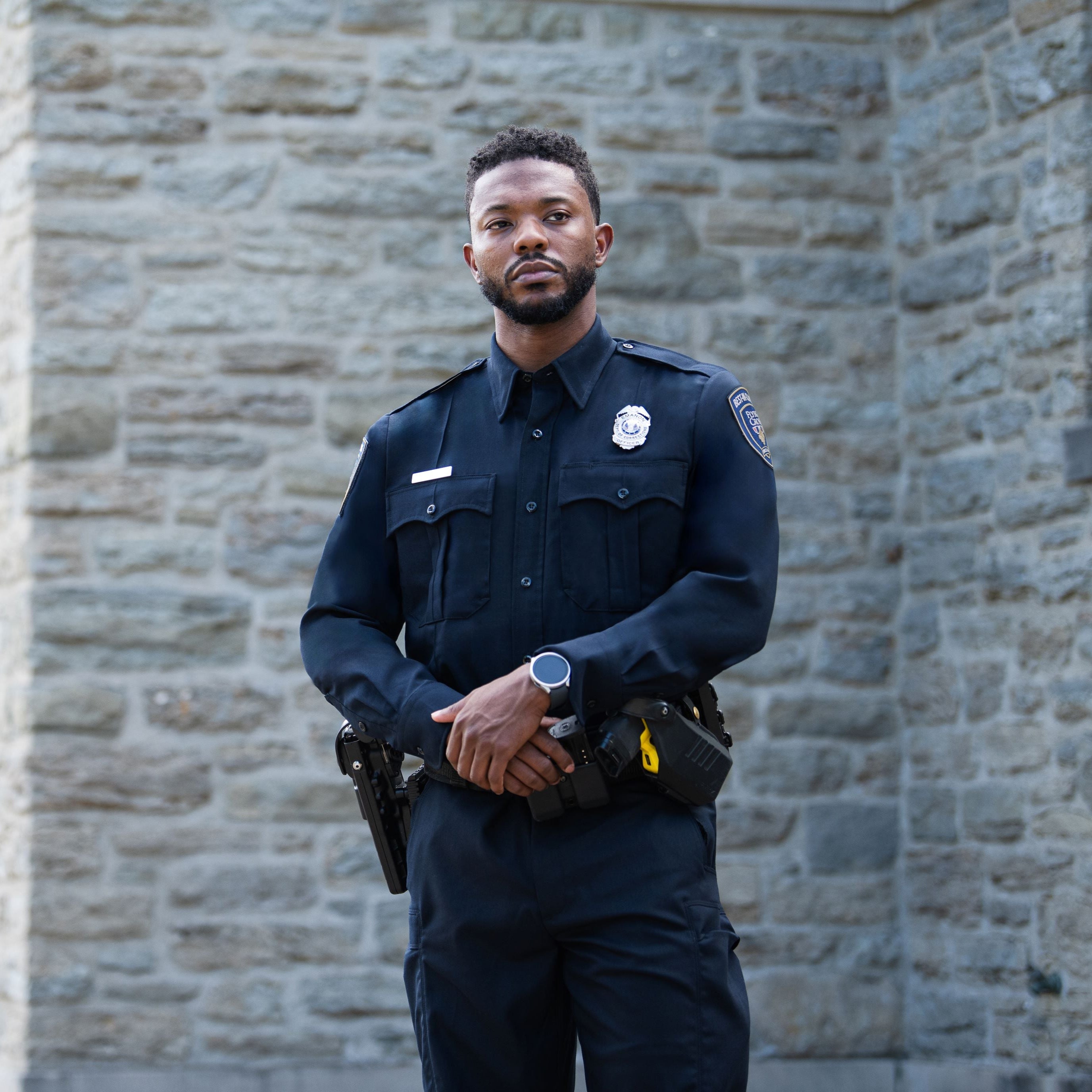 Photograph of a male police officer in front of a rustic stone wall wearing a dark blue uniform a silver smartwatch and a yellow detail on his utility belt