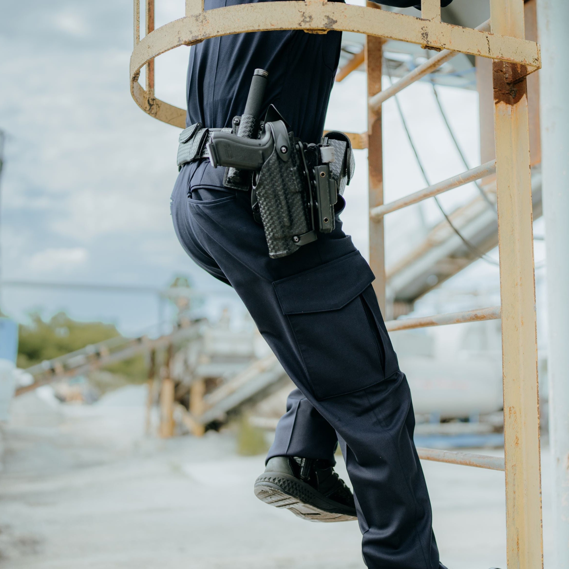 Photograph of a security guard climbing a rusty metal ladder, showcasing a black firearm in a textured holster, dark blue uniform pants with cargo pockets, and worn black tactical boots against a blurred industrial backdrop.