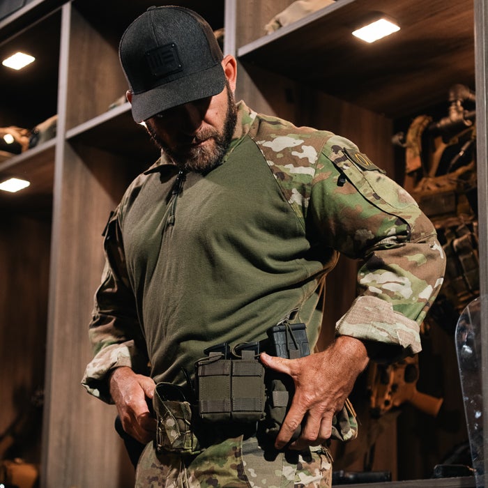 Photography of a man in camouflage, adjusting his tactical gear in a dimly lit room with wooden shelves and firearms visible featuring a dark green shirt, a black hat, and olive drab pouches