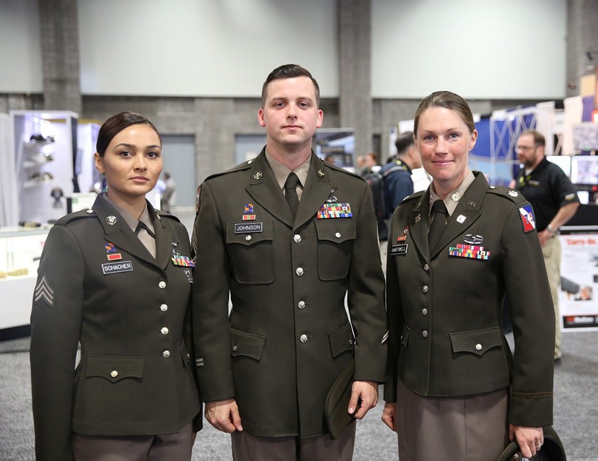 Photograph three US Army soldiers in olive green service uniforms at an indoor expo featuring various colorful ribbons medals and name tags