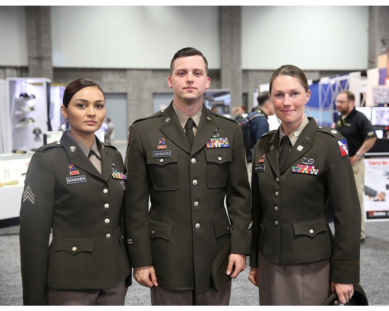Photograph three US Army soldiers in olive-green service uniforms at a trade show featuring colorful rank insignia, name tapes, and a variety of exhibitors in the background