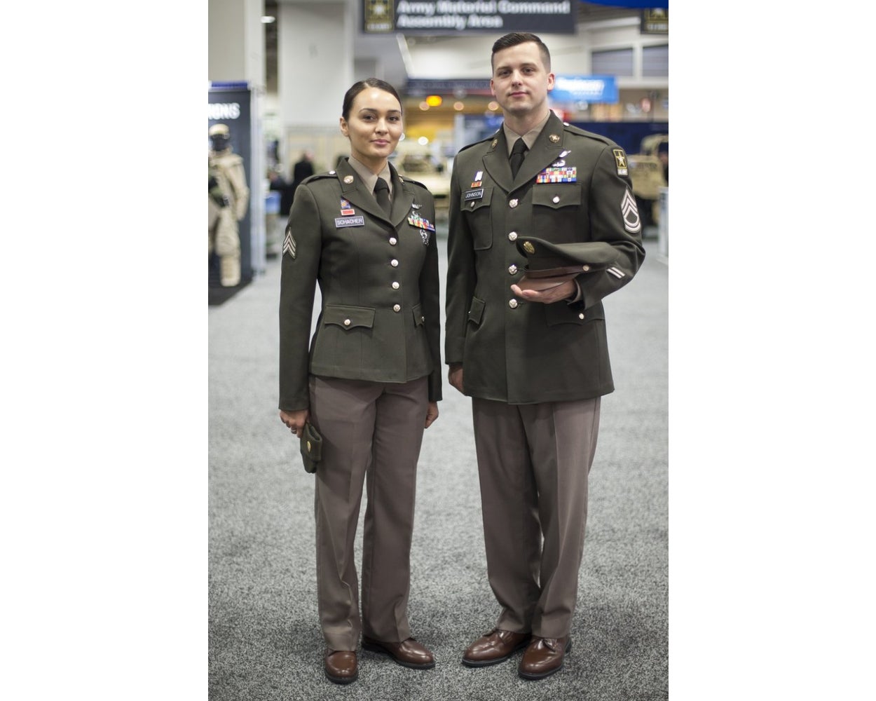 Photograph of two Army sergeants in olive green service uniforms with brown shoes and nametags at an Army Material Command event featuring army vehicles and signage in the background