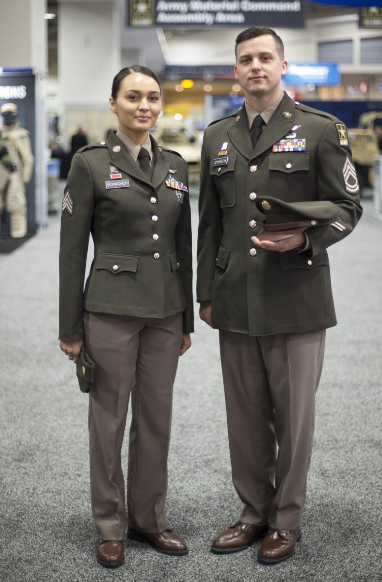 Photograph two Army soldiers in olive green service uniforms with name tapes and rank insignia at an indoor event featuring muted colors and brown leather shoes against a backdrop of exhibit booths and signage