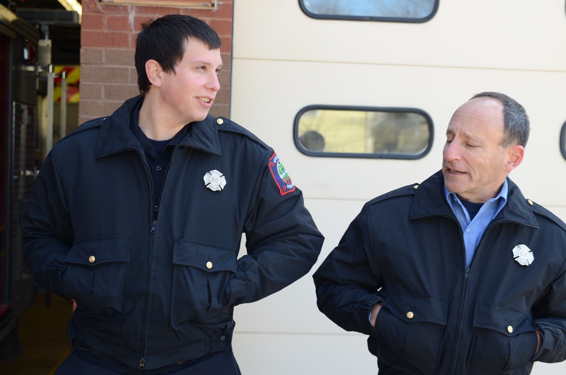 Photograph two firefighters in dark jackets with gold buttons and patches outside a fire station featuring a red brick wall and garage doors