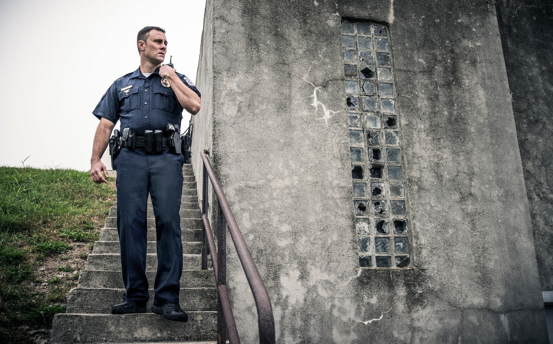 Photograph of a police officer using a radio on a concrete staircase next to a weathered building with a damaged, gridded window and rusty handrail showing the officer's dark uniform, tactical gear, and serious expression