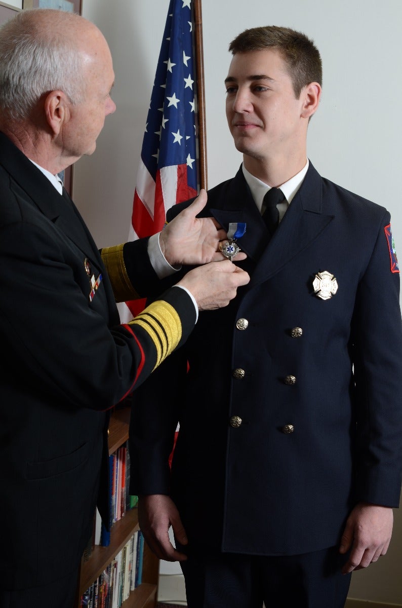 Photograph of a firefighter receiving a medal near an American flag and a bookshelf with colorful books in an office setting, the medal is silver with blue ribbon, the firefighter's uniform has gold buttons and a patch