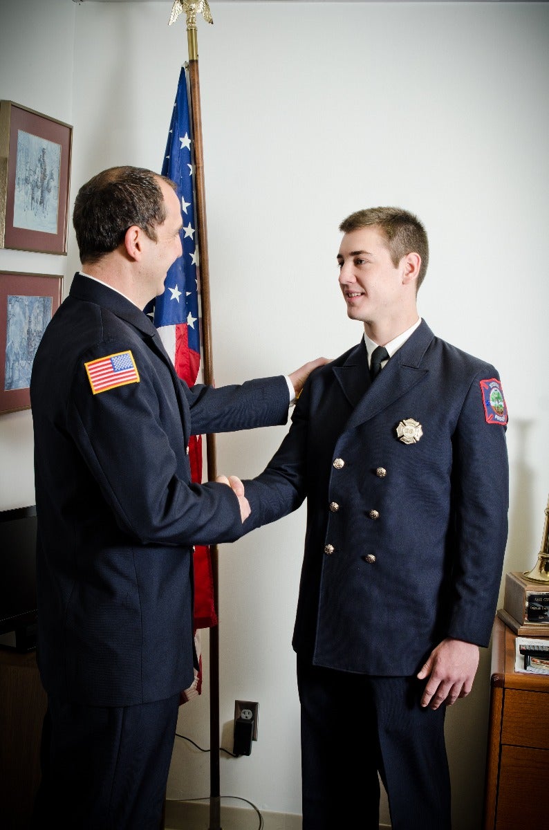 Photograph two firefighters shaking hands in an office near an American flag with gold buttons a firefighter badge and framed artwork