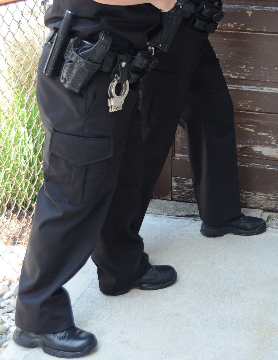 Photograph showing a police officer's lower body near a weathered wooden door and chain link fence with black tactical boots, shiny handcuffs, and a black firearm in a holster.