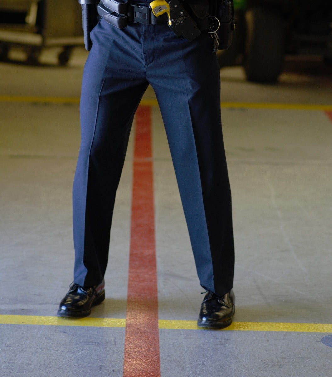 Photograph of a police officer's legs and feet in a garage with yellow and red floor markings, showcasing black polished shoes, a dark-blue uniform, and a utility belt with a yellow detail.