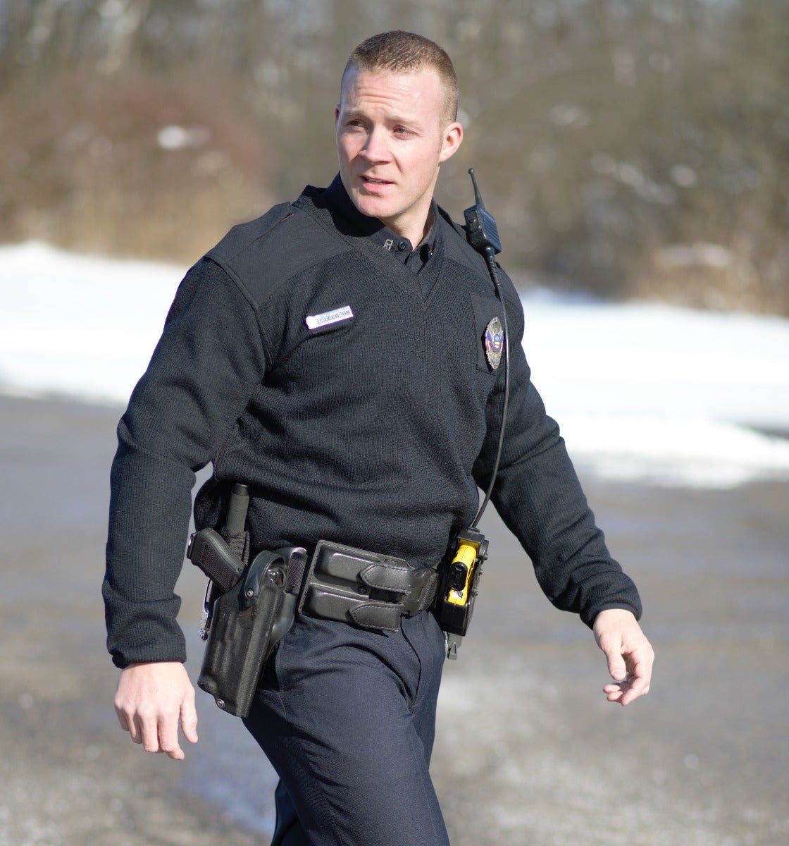 Photograph of a police officer in a black uniform patrolling a snowy, wintry roadside with a handgun, yellow taser, and name tag visible