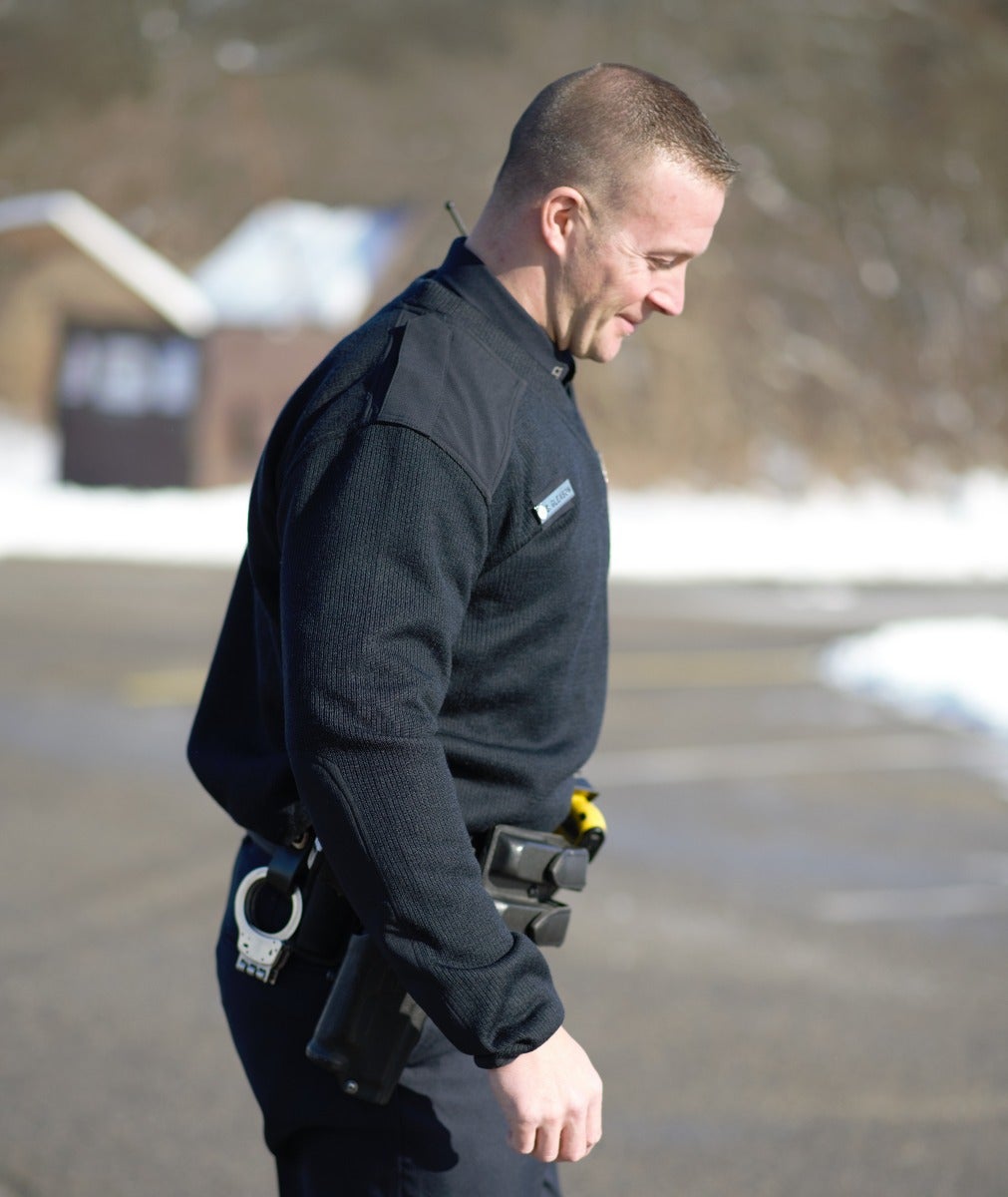 Photograph of a police officer in a dark ribbed sweater and duty belt with handcuffs and pepper spray walking outside near a building with a snowy background