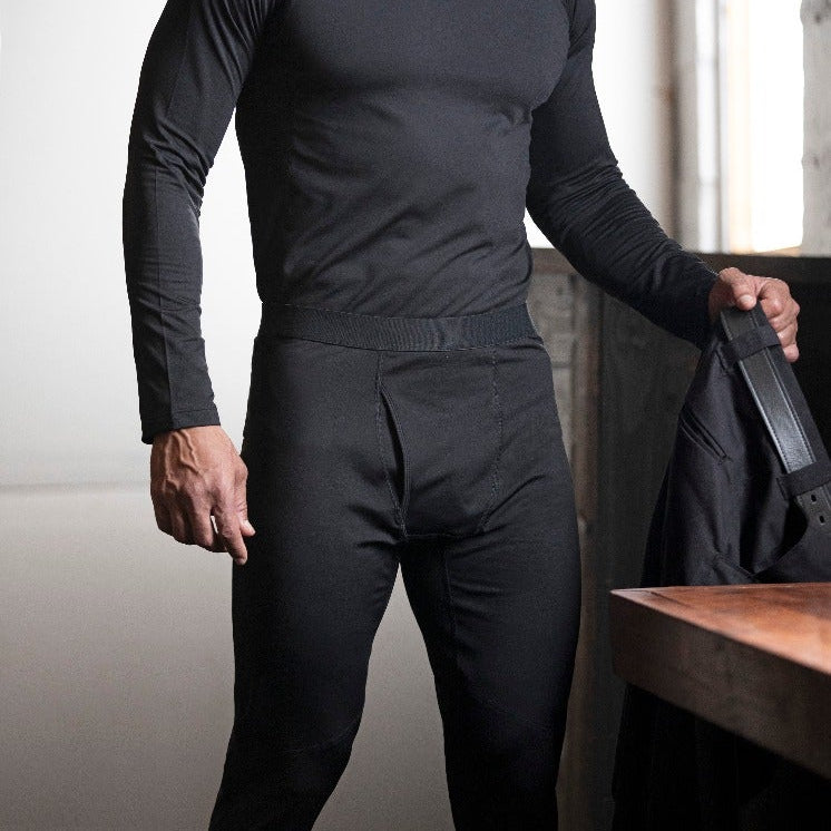 Product shot of a man indoors modeling black thermal underwear with a ribbed waistband and a dark wood table in the background