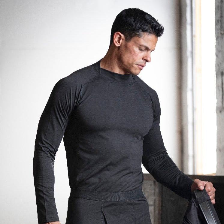 Photograph model wearing a black mock-neck thermal shirt and matching pants in a minimalist industrial setting with high ceilings and muted light