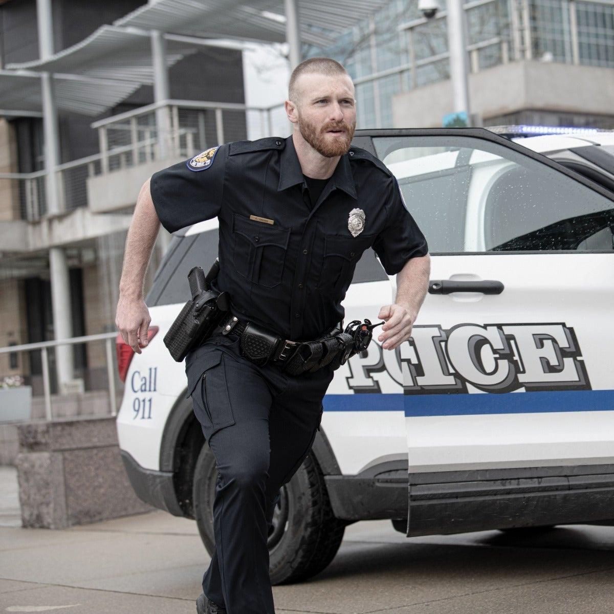 Photograph Police officer running towards a police car near modern buildings a black gun holster a police badge and a dark blue stripe on the car