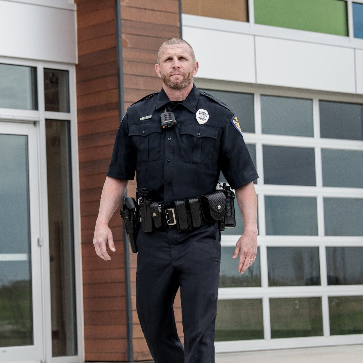 Photograph of a police officer in uniform walking outside a modern building with wood paneling, large glass doors, and a white paneled garage door featuring a black utility belt, a radio, and a police badge
