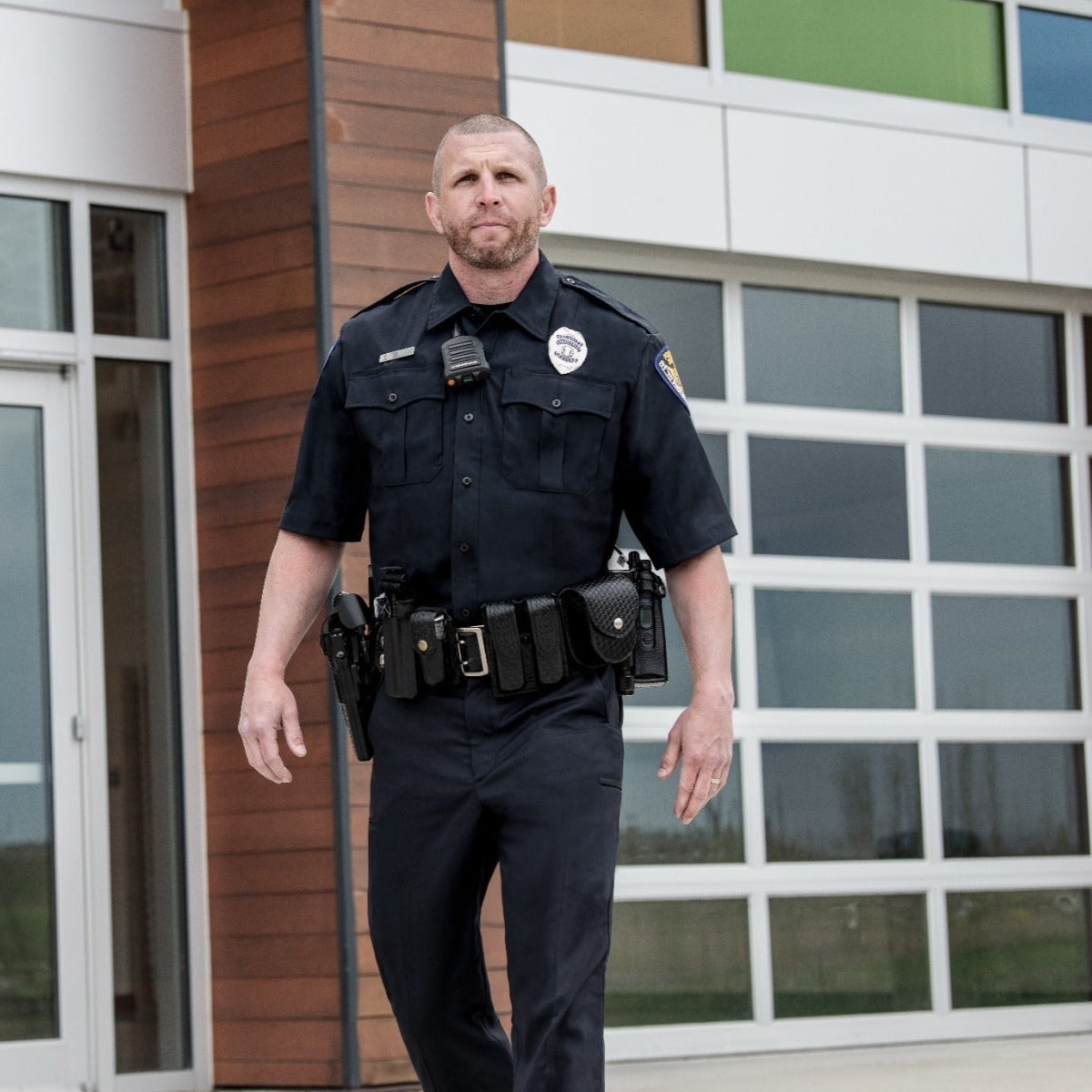 Photograph of a police officer in front of a modern building with large windows and wood paneling, wearing a dark uniform with a radio and utility belt, showcasing his badge and a serious expression