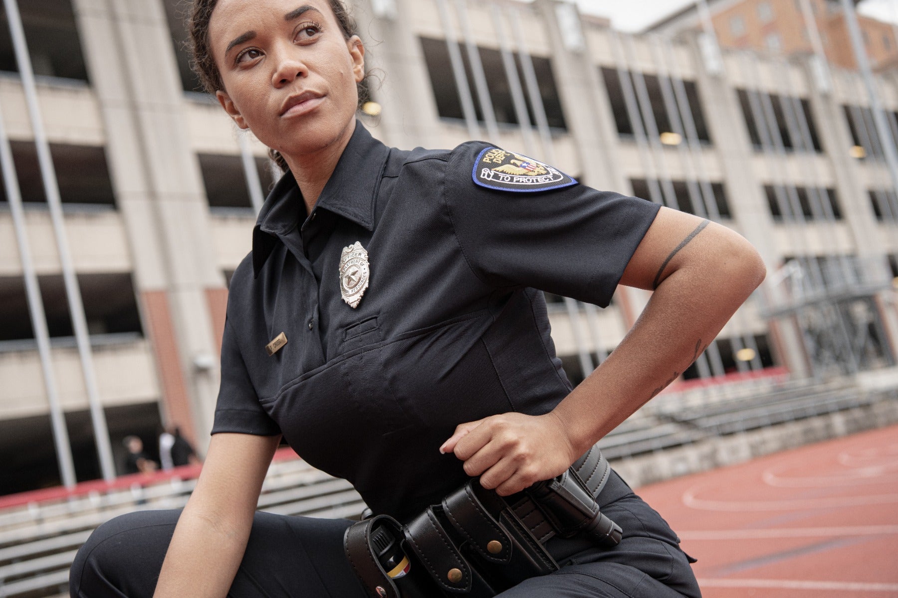 Photograph female police officer in a black uniform at an outdoor urban sports facility near a multi-story parking garage with a silver badge, black leather holster and a small forearm tattoo