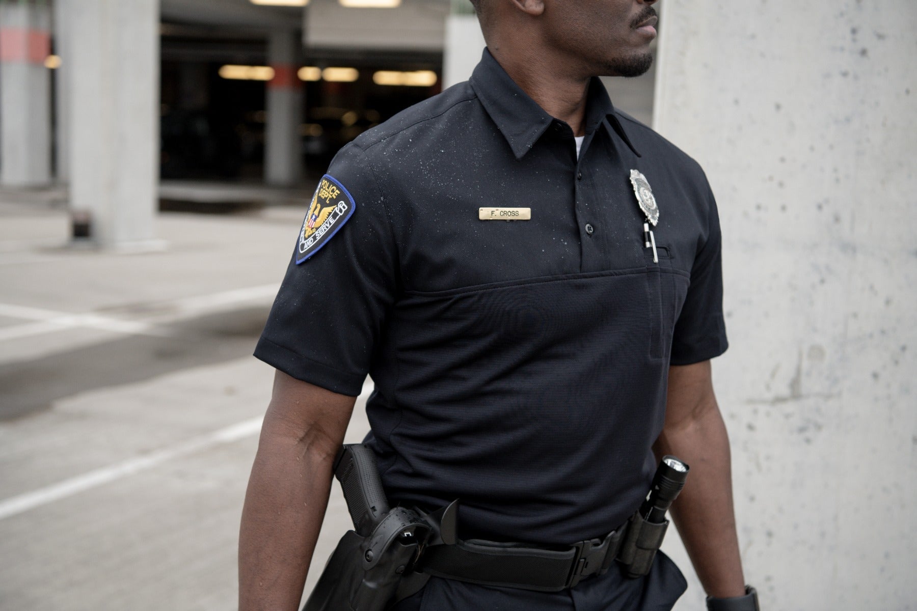 Photograph of a police officer in a parking garage wearing a black uniform with a gold nameplate and a detailed shoulder patch showing an eagle and the words Police Dept and To Serve and Protect featuring a handgun and flashlight on his hip