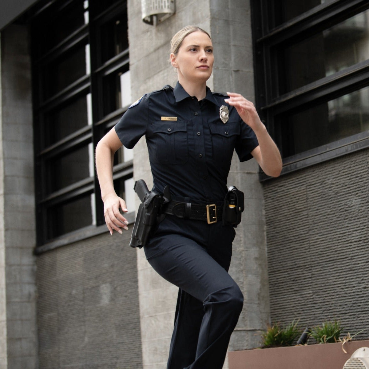 Photograph of a female police officer in uniform running outside a modern building with large windows and textured grey walls, she carries a gun and her badge is visible, her expression is serious and determined, and her hair is neatly pulled back.