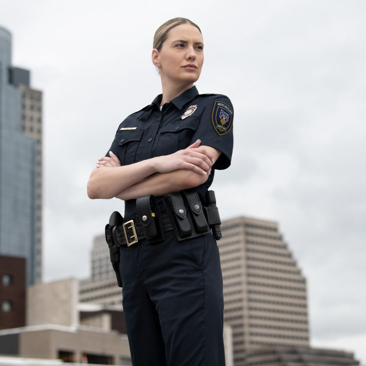Photograph of a female police officer with arms crossed in a city setting featuring high-rise buildings and a gold belt buckle with a dark blue uniform and a Best in Class patch