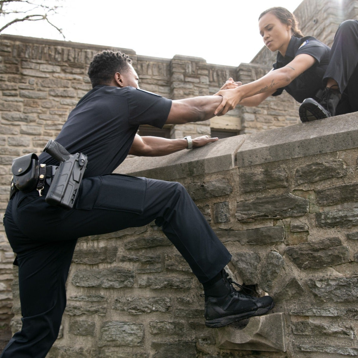 Photograph two police officers assisting each other over a stone wall near a historic building featuring weathered stonework and intricate details a watch and a tattoo are visible.