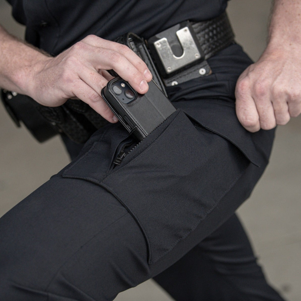 Photograph showing a person placing a smartphone in a zippered tactical pants pocket featuring a black leather phone case and a metallic police belt buckle