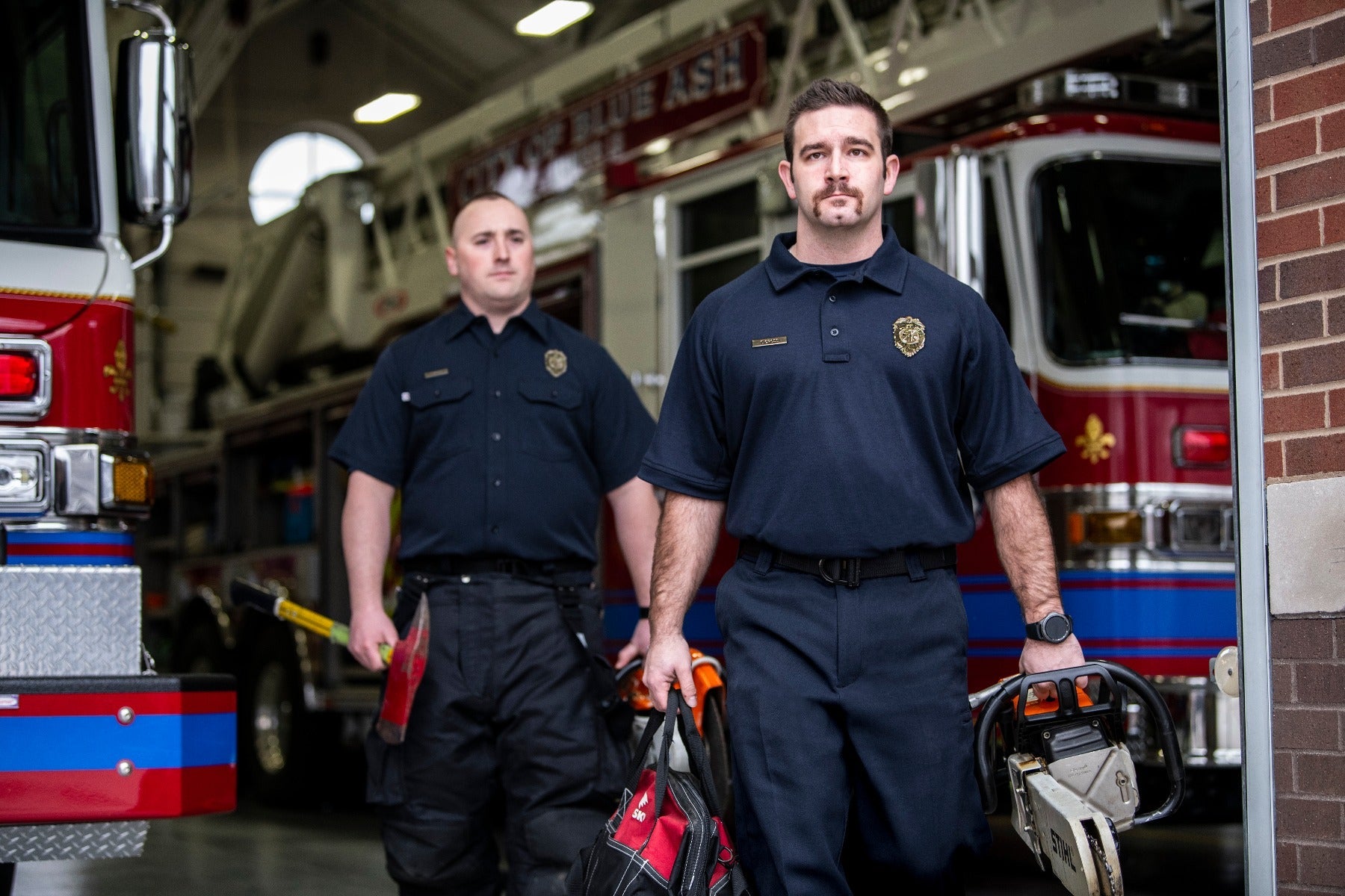 Photograph of two firefighters in a fire station carrying equipment including an axe and chainsaw in front of a red firetruck with blue and gold details and a fleur-de-lis emblem