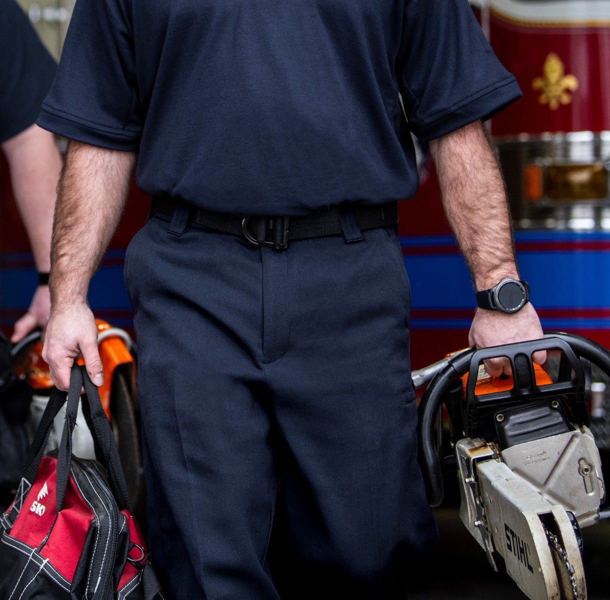 Photograph firefighter carrying a Stihl chainsaw and a red bag near a firetruck with blue and red stripes and a fleur-de-lis emblem showing a black tactical belt and a smartwatch