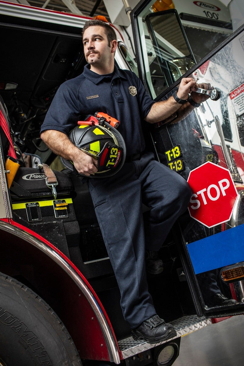 Professional photograph of a firefighter in a fire station holding a helmet with a T-13 marking near a Pierce 100 firetruck featuring a stop sign and bright yellow and red details