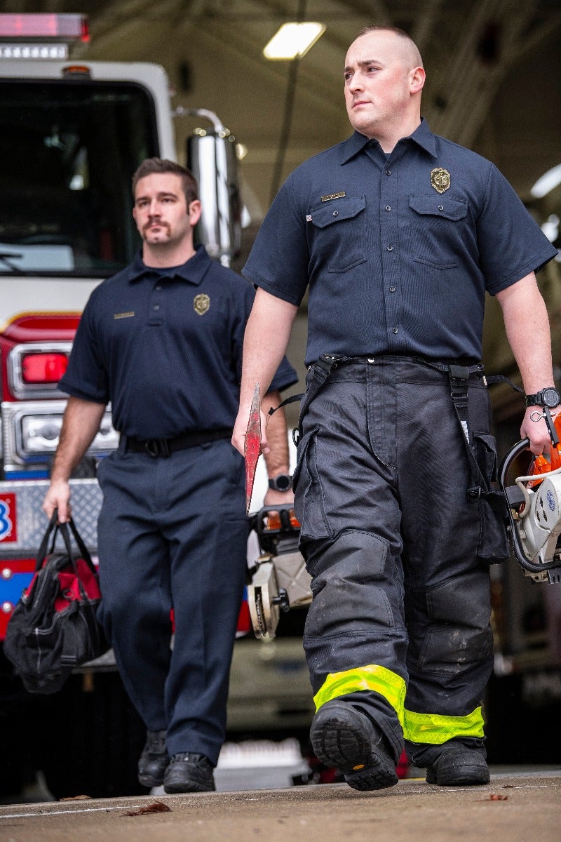 Photograph two firefighters in a fire station near a firetruck carrying equipment including a chainsaw and an axe featuring a vibrant yellow reflective stripe on their pants and dark navy uniforms