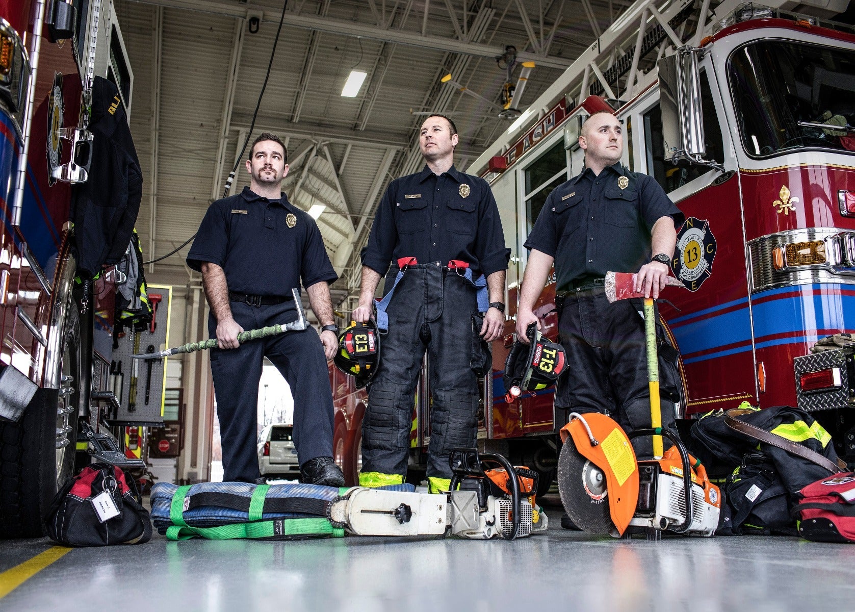 Photograph three firefighters in a fire station with a red firetruck featuring a fleur-de-lis emblem and blue and white stripes holding rescue tools including a chainsaw and a circular saw next to green and blue rescue equipment